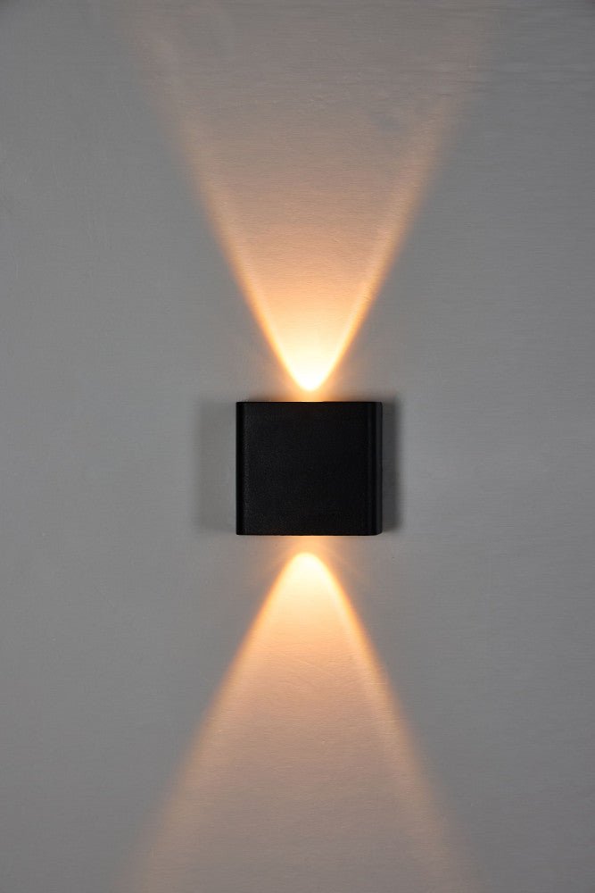 Zwart wandlamp met warm wit licht, geïnstalleerd op een grijze muur boven een donkere bank. Geschikt voor buitenverlichting en avondelijke sfeercreatie in tuinen of terrassen.
