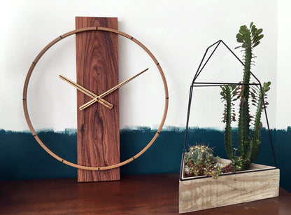 Een moderne wandklok met een goudkleurige metalen ring en een houten paneel met donkerbruine, gestreepte textuur. De dunne wijzers en het merk 'NEXTIME' zijn zichtbaar. Ideaal voor minimalistische interieurs en moderne woonkamers.