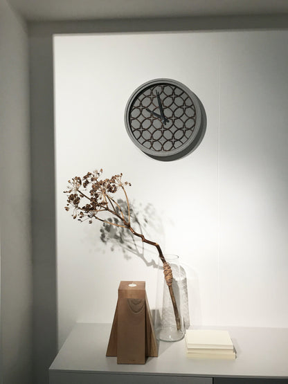 Een moderne wandklok met grijze betonrand en houten geometrisch patroon. Zilverkleurige wijzers en het merk 'NEXTIME' zijn zichtbaar. Ideaal voor minimalistische interieurs of als decoratief element in woon- en werkkamers.