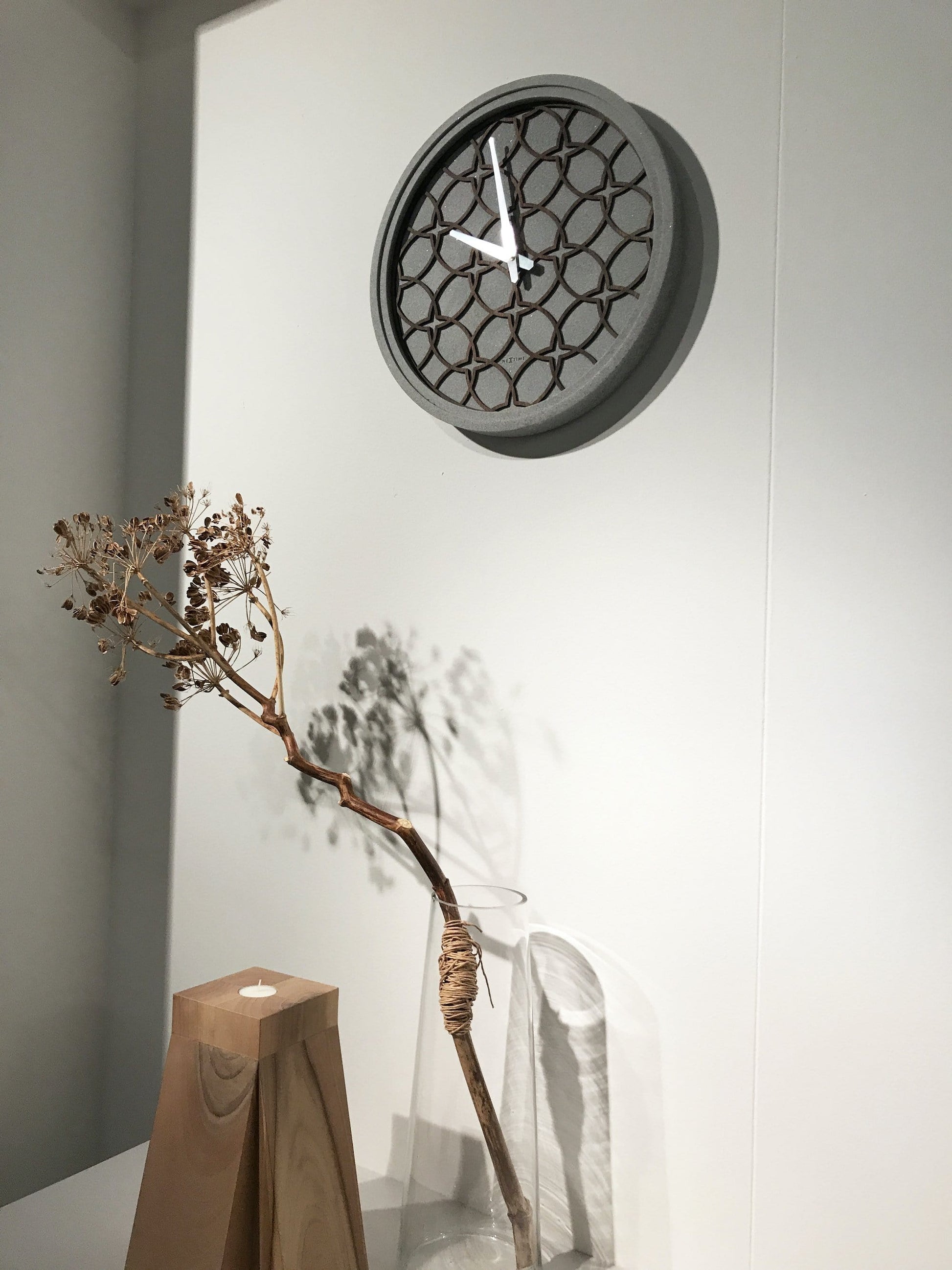 Een moderne wandklok met een grijze betonrand en een houten, geometrisch patroon. Zilverkleurige wijzers en het merk 'NEXTIME' zijn zichtbaar. Geschikt voor minimalistische interieurs en als decoratief element in woon- of kantoorruimtes.