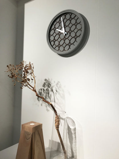Een moderne wandklok met een grijze betonrand en een houten, geometrisch patroon. Zilverkleurige wijzers en het merk 'NEXTIME' zijn zichtbaar. Geschikt voor minimalistische interieurs en als decoratief element in woon- of kantoorruimtes.