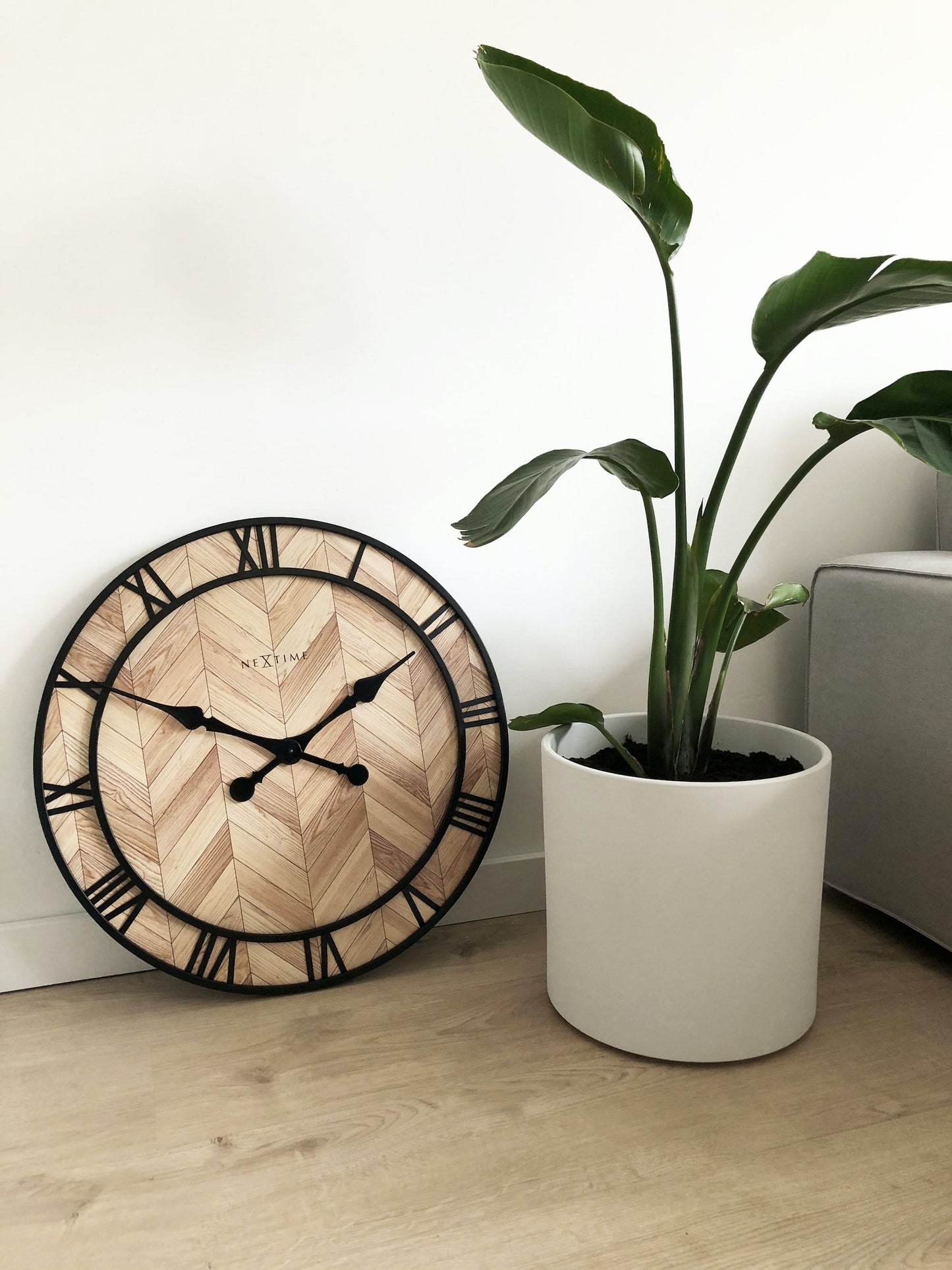 Een ronde wandklok met houten chevronpatroon en zwarte Romeinse cijfers. De klok heeft een zwart metalen frame en wijzers, geschikt voor moderne interieurs of als decoratief element in een woonkamer.