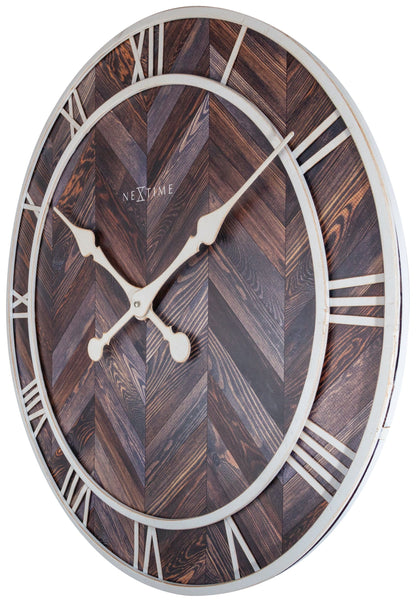 Een ronde wandklok met houten chevronpatroon en witte Romeinse cijfers. De witte wijzers en 'NEXTIME' merknaam zijn duidelijk zichtbaar. Perfect voor een moderne woonkamer of kantoordecoratie.