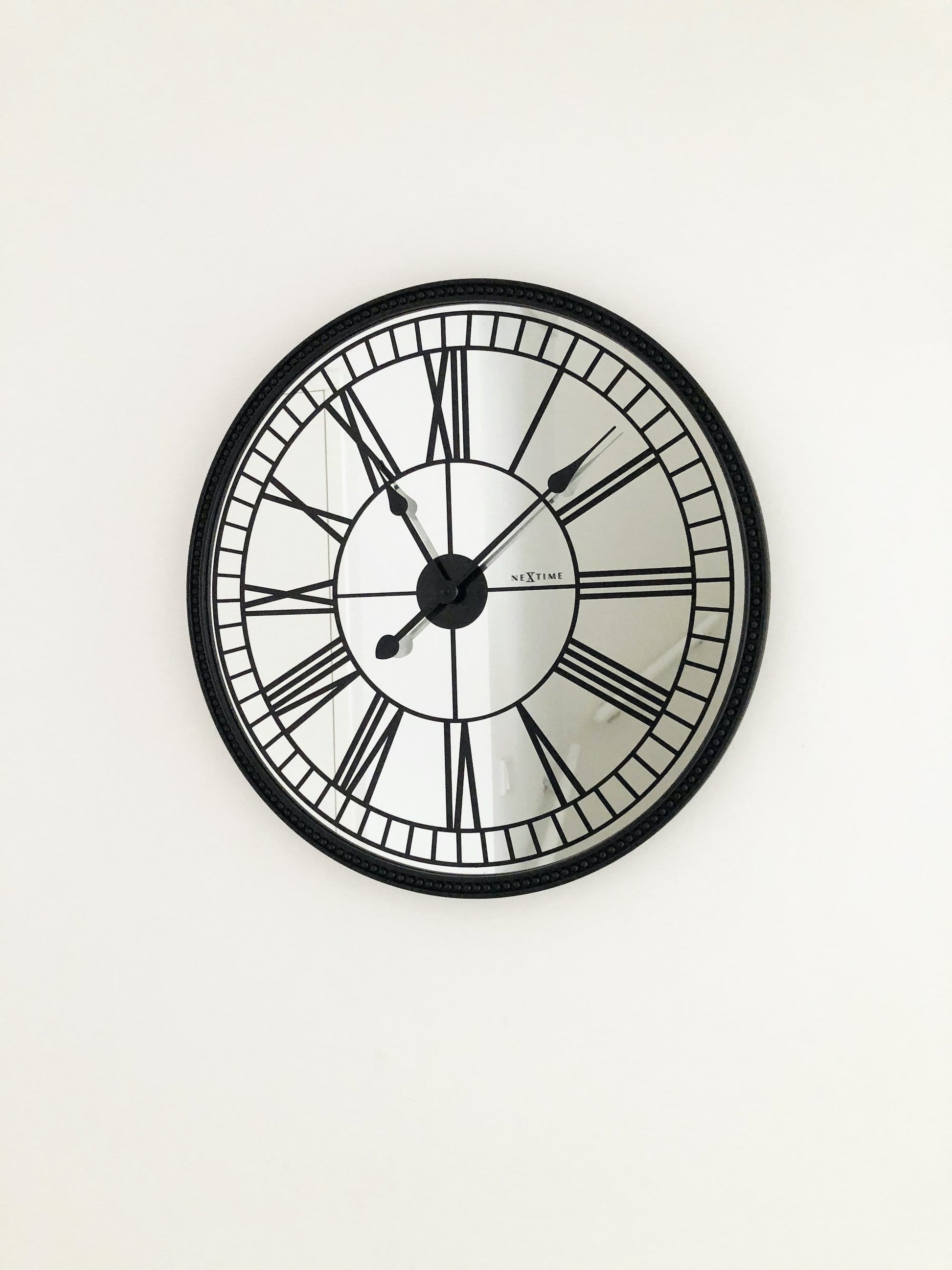 Een klassieke wandklok met zwart metalen kader en witte wijzerplaat met Romeinse cijfers. De wijzers en het merk 'NEXTIME' zijn duidelijk zichtbaar. Geschikt voor woonkamer of kantoordecoratie.