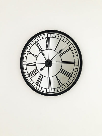 Een klassieke wandklok met zwart metalen kader en witte wijzerplaat met Romeinse cijfers. De wijzers en het merk 'NEXTIME' zijn duidelijk zichtbaar. Geschikt voor woonkamer of kantoordecoratie.