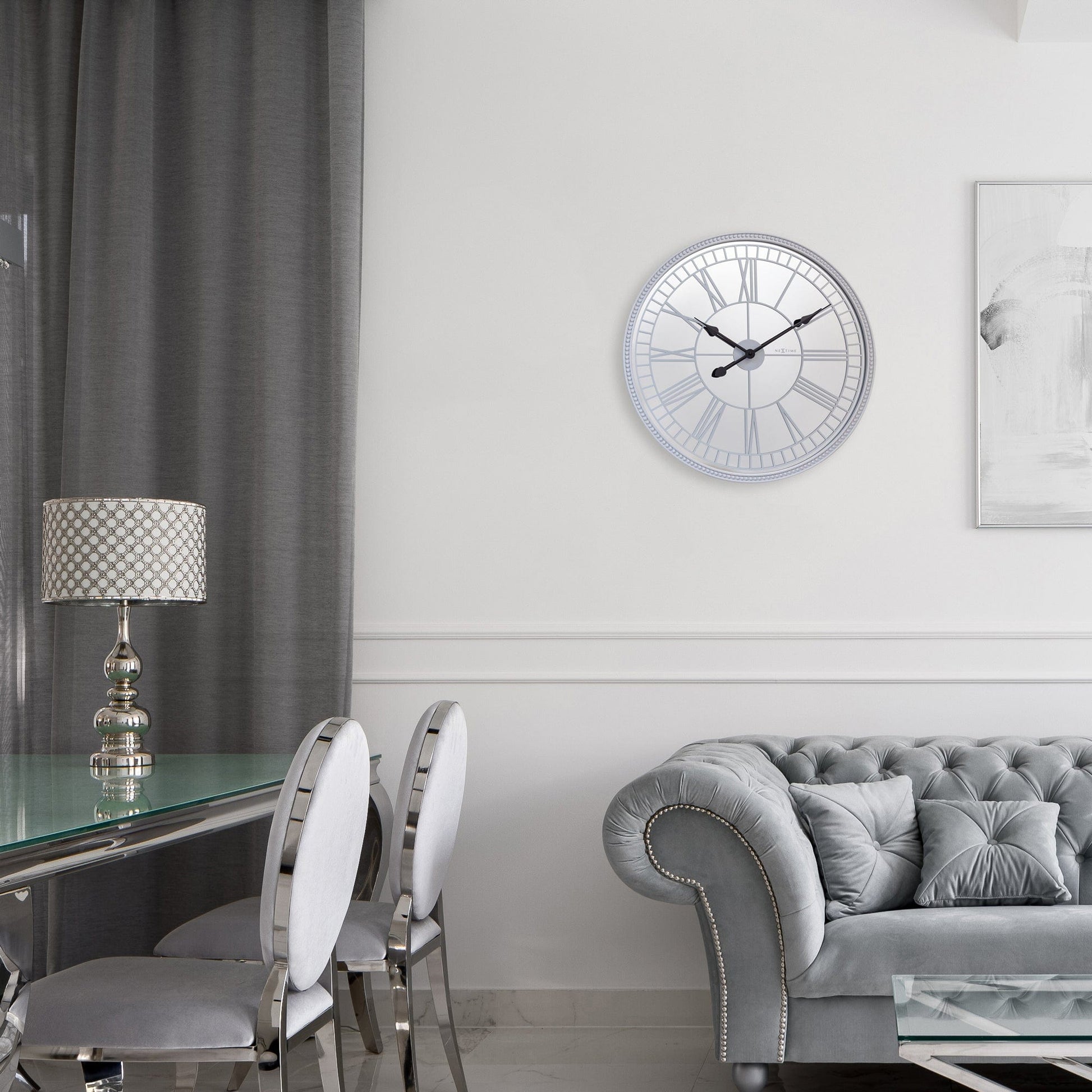 Een elegante, zilverkleurige wandklok met Romeinse cijfers en zwarte wijzers. De klassieke klok heeft een glanzende rand met bolletjes en is ideaal voor een moderne woonkamer of kantoor.