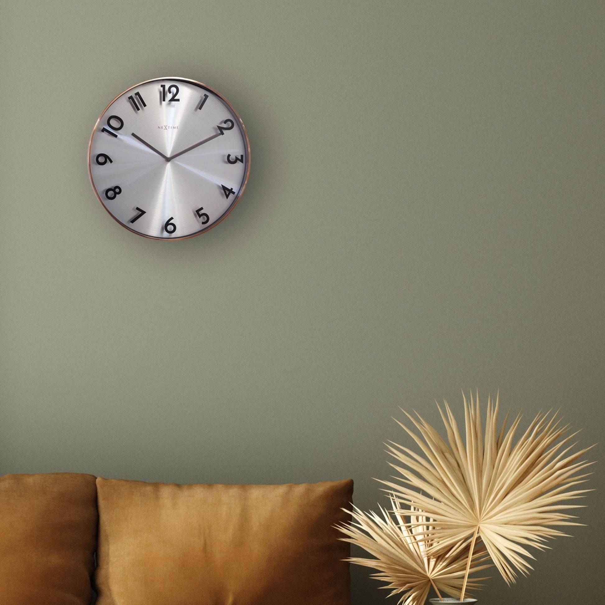 Een moderne wandklok met zilverkleurig, geborsteld wijzerplaat en zwarte cijfers, omlijst met een warme koperen rand. Geschikt voor minimalistische interieurs en als decoratief element in woon- of kantoorruimtes.