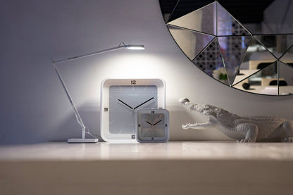 Een minimalistische, zilverkleurige wandklok met zwarte cijfers en wijzers, in een witte kader. Geschikt voor moderne interieurs en als decoratief element in woon- of kantoorruimtes.