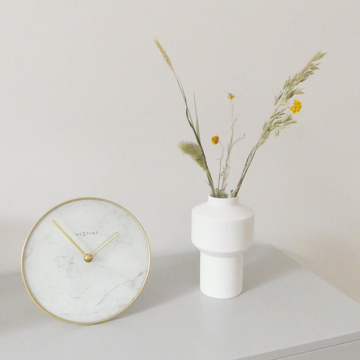 Een ronde wandklok met wit marmeren oppervlak en goudkleurige wijzers. De klok heeft een goudkleurige rand en het merk 'NEXTIME' in het midden, geschikt voor moderne interieurs.