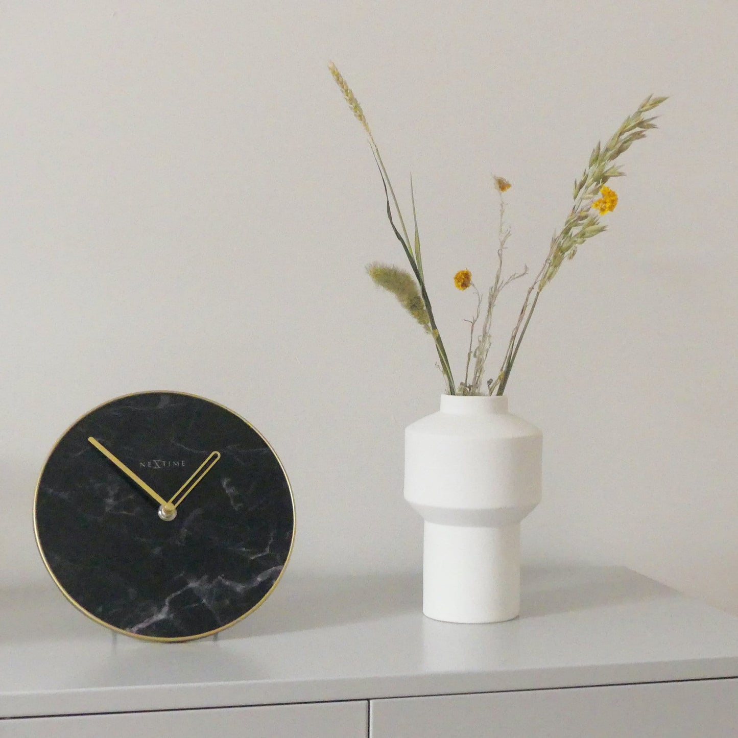 Een elegante wandklok met zwart marmeren wijzerplaat en goudkleurige wijzers. De minimalistische klok heeft een goudkleurige rand en het merk NEXtime, ideaal voor moderne interieurs en als decoratief element in woon- of kantoorruimtes.