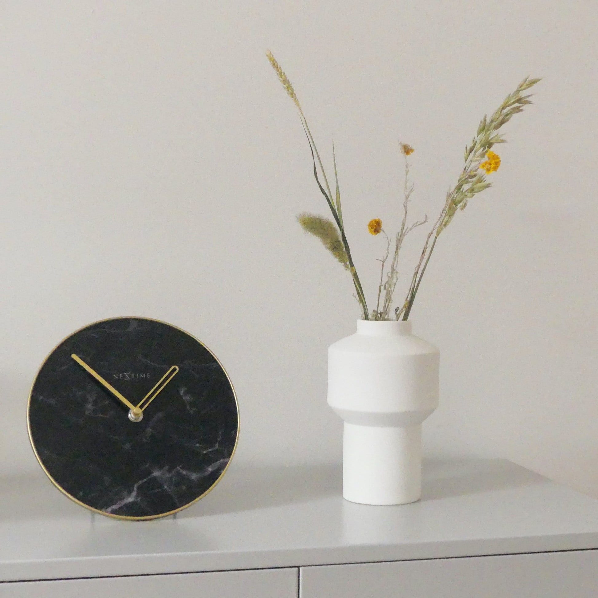 Een elegante wandklok met zwart marmeren wijzerplaat en goudkleurige wijzers. De minimalistische klok heeft een goudkleurige rand en het merk NEXtime, ideaal voor moderne interieurs en als decoratief element in woon- of kantoorruimtes.