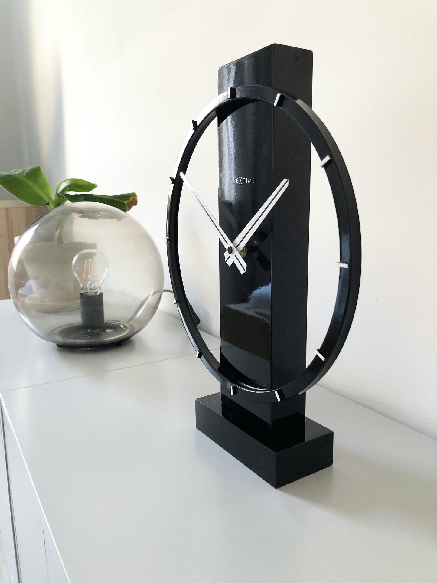 Een moderne, zwarte tafelklok van het merk Nextime met een minimalistisch ontwerp, witte wijzers en een glanzende, cirkelvormige rand. Perfect voor een stijlvolle woonkamer of kantoordecoratie.