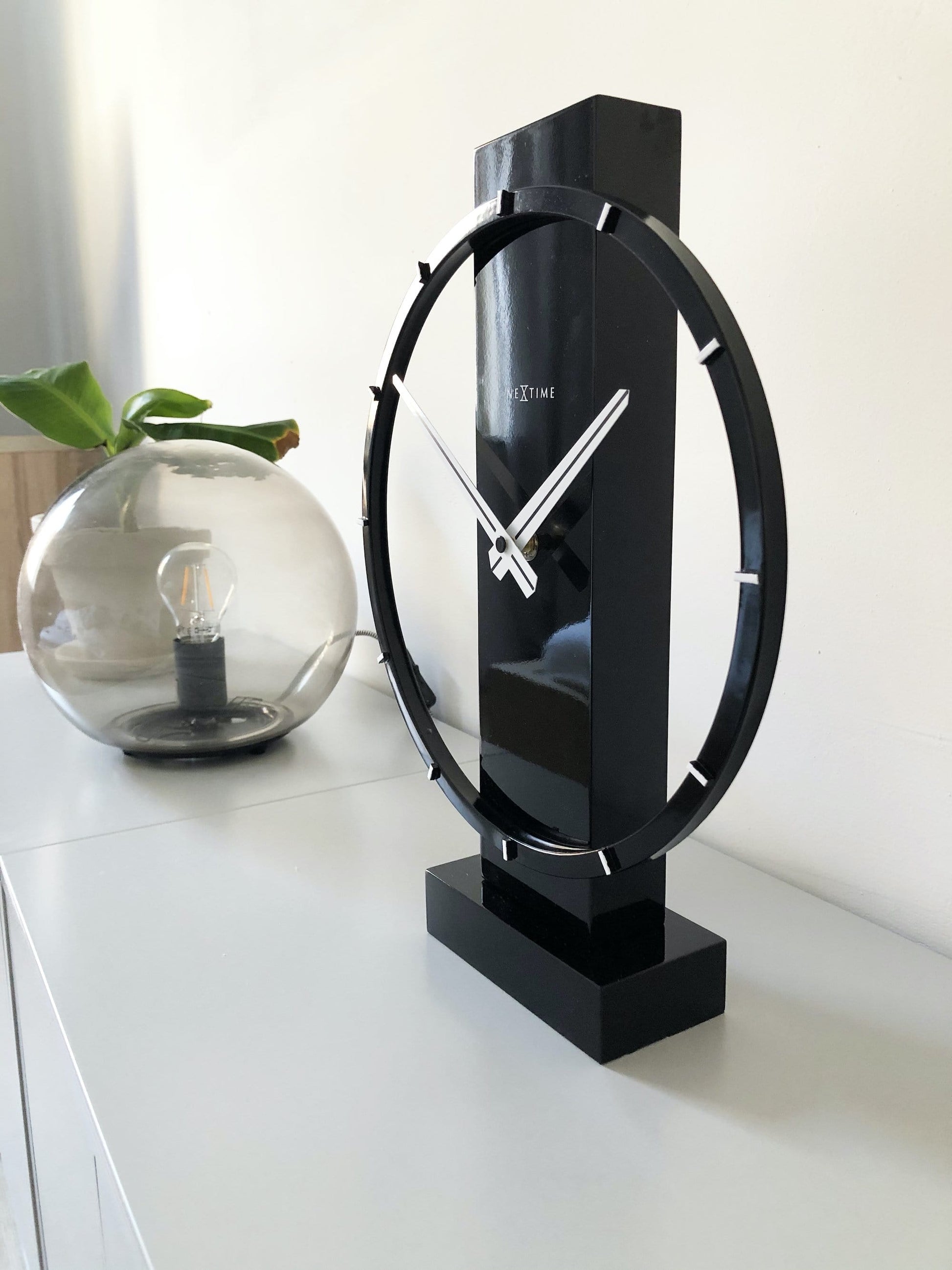 Een moderne, zwarte tafelklok van het merk Nextime met een minimalistisch ontwerp, witte wijzers en een glanzende, cirkelvormige rand. Perfect voor een stijlvolle woonkamer of kantoordecoratie.