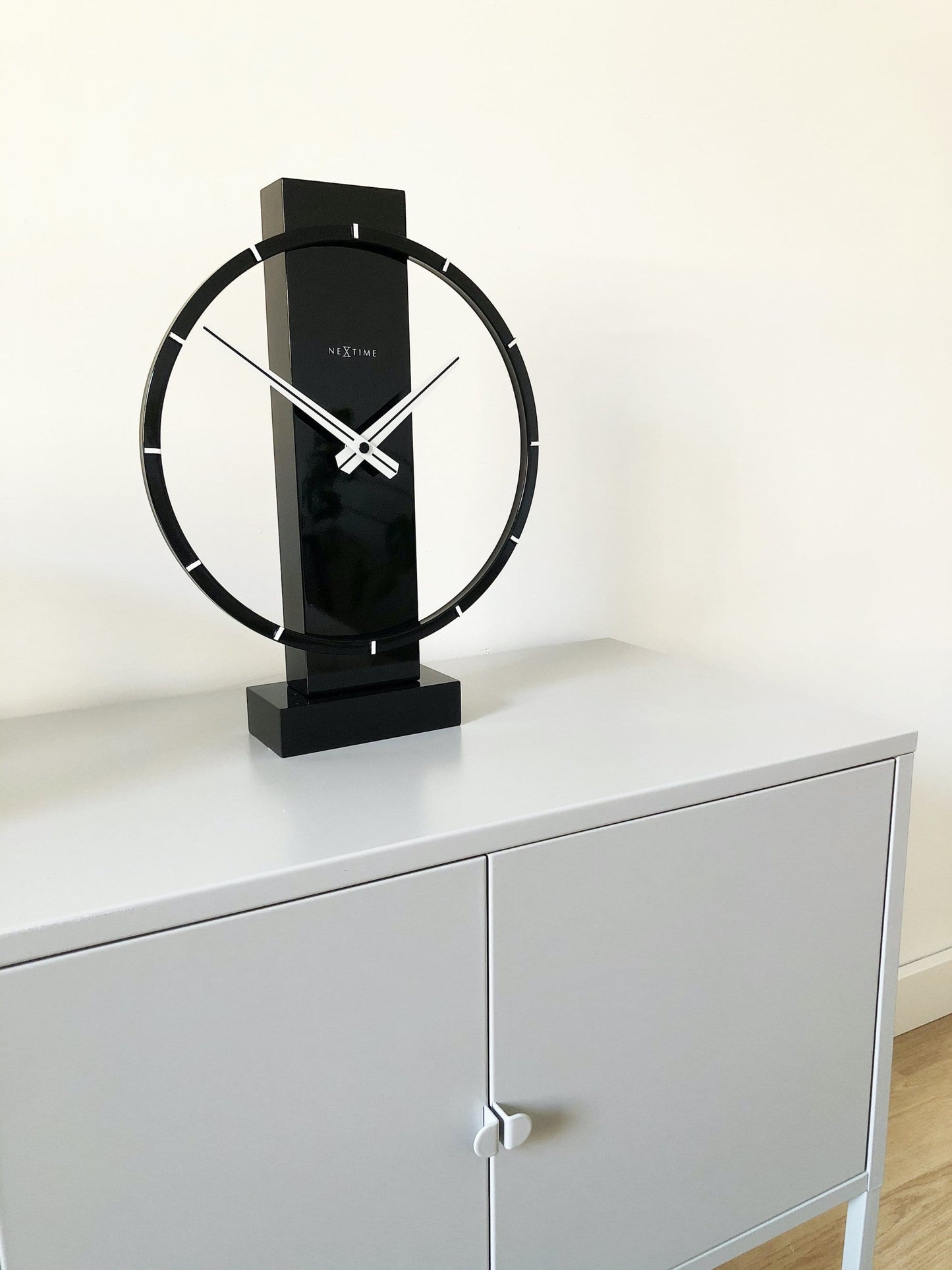 Een moderne, zwarte tafelklok van het merk Nextime met een minimalistisch ontwerp, witte wijzers en een glanzende, cirkelvormige rand. Ideaal voor een stijlvolle woonkamer of kantoordecoratie.