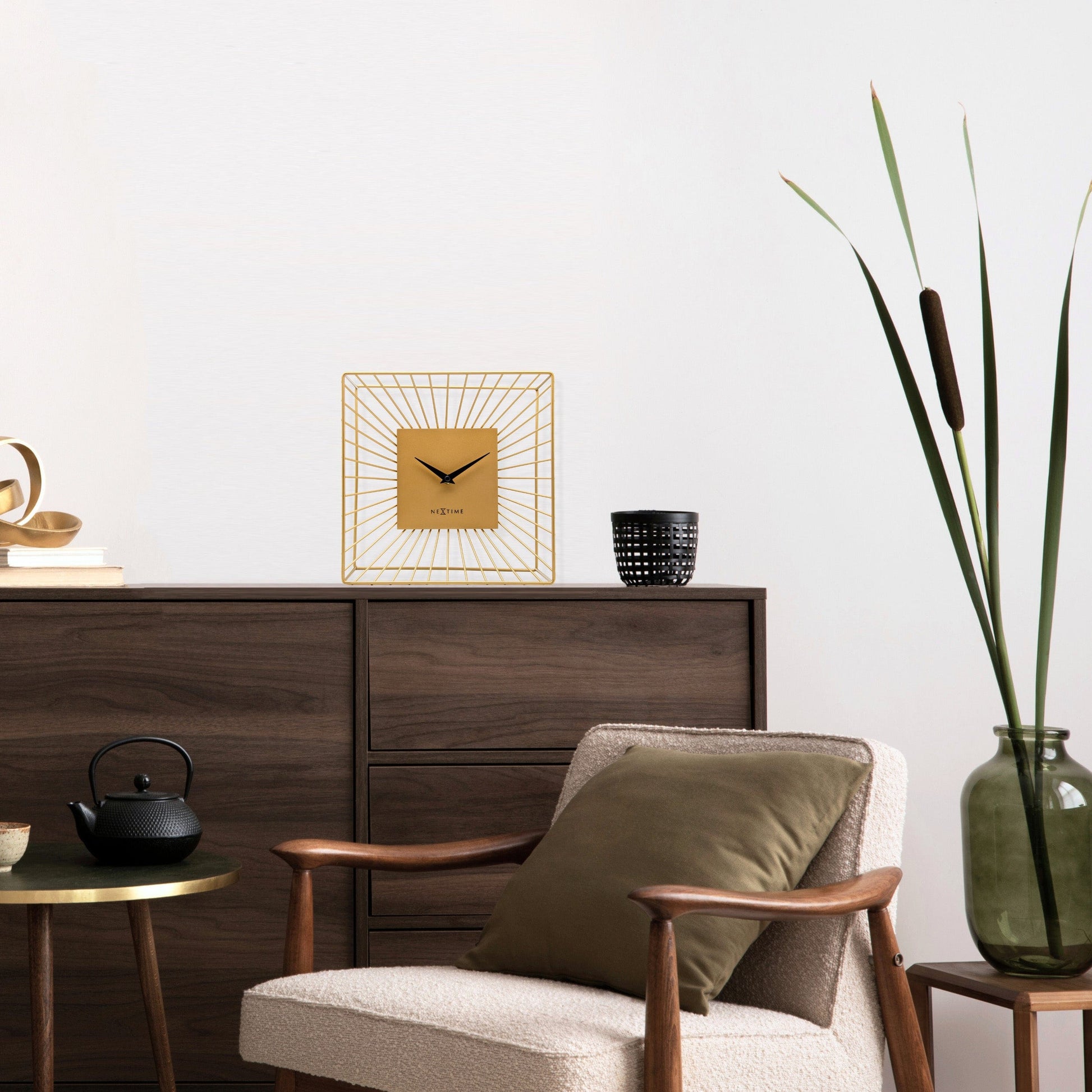 Een goudkleurige wandklok met een minimalistisch ontwerp, een vierkante kast van metaal met stralende lijnen en zwarte wijzers. Geschikt voor moderne interieurs en als decoratief element in woon- of kantoorruimtes.
