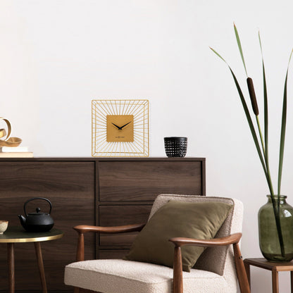 Een goudkleurige wandklok met een minimalistisch ontwerp, een vierkante kast van metaal met stralende lijnen en zwarte wijzers. Geschikt voor moderne interieurs en als decoratief element in woon- of kantoorruimtes.