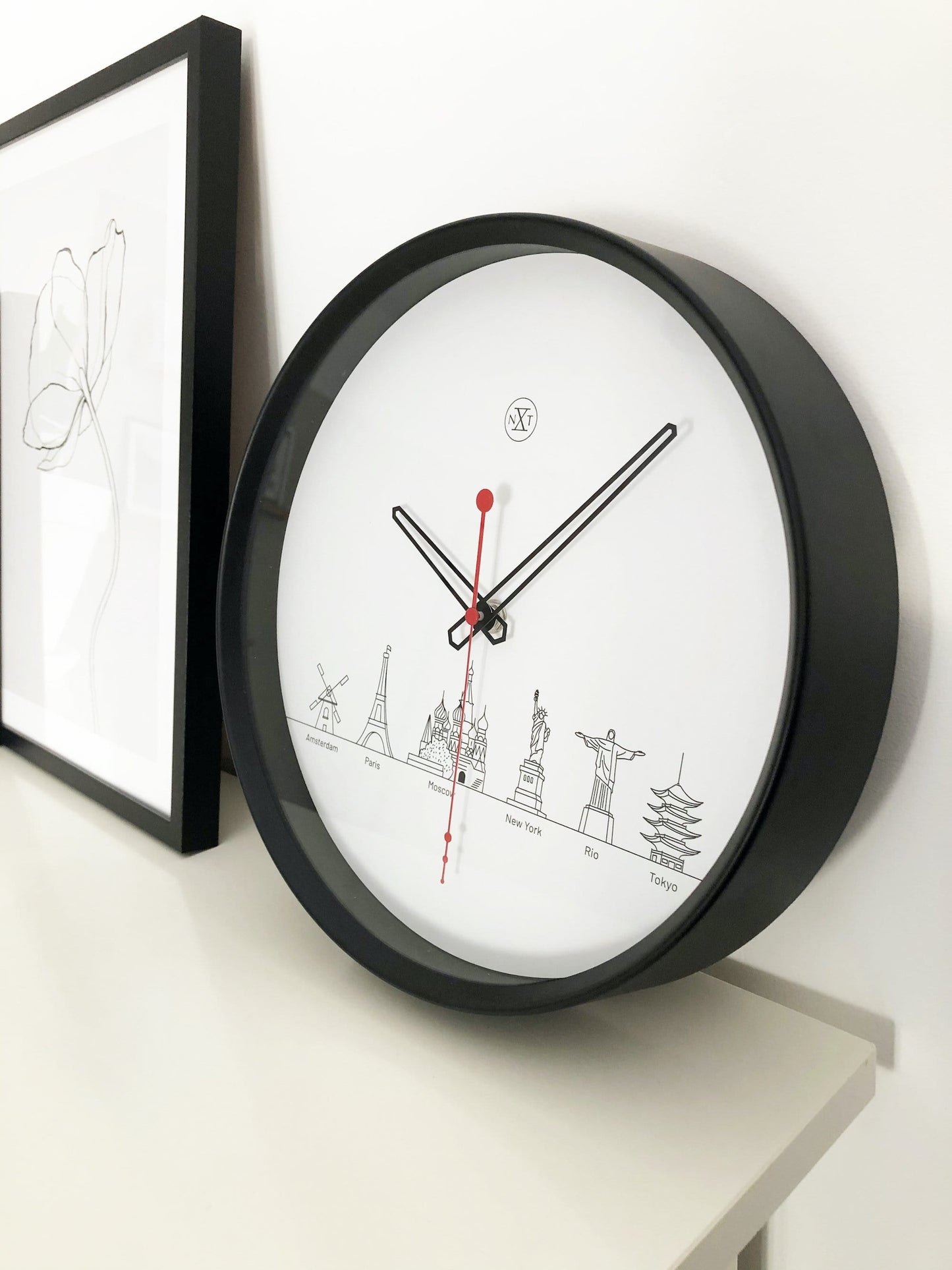Een minimalistische wandklok met zwart kader en wit oppervlak, voorzien van zwart wijzerplaat en rode secondewijzer. De wijzerplaat toont gestileerde wereldstadiconen, ideaal voor een moderne, wereldwijde interieurdecoratie.