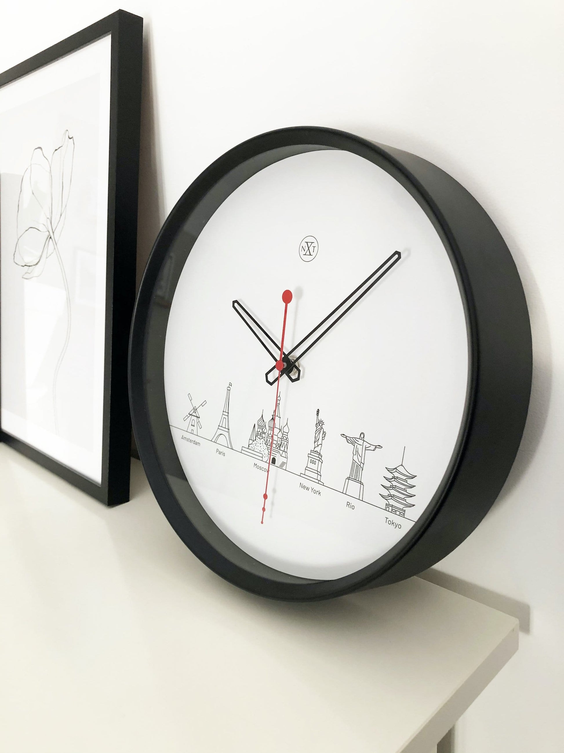 Een minimalistische wandklok met zwart kader en wit oppervlak, voorzien van zwart wijzerplaat en rode secondewijzer. De wijzerplaat toont gestileerde wereldstadiconen, ideaal voor een moderne, wereldwijde interieurdecoratie.
