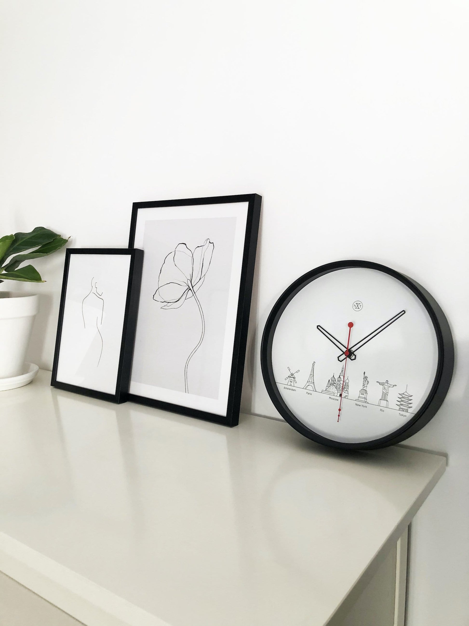 Een minimalistische wandklok met zwart kader en witte wijzerplaat, voorzien van zwart en rood wijzers en silhouetten van wereldstadsmarkeringen. Ideaal voor een moderne woonkamer of kantoorruimte met een reis- of wereldthema.