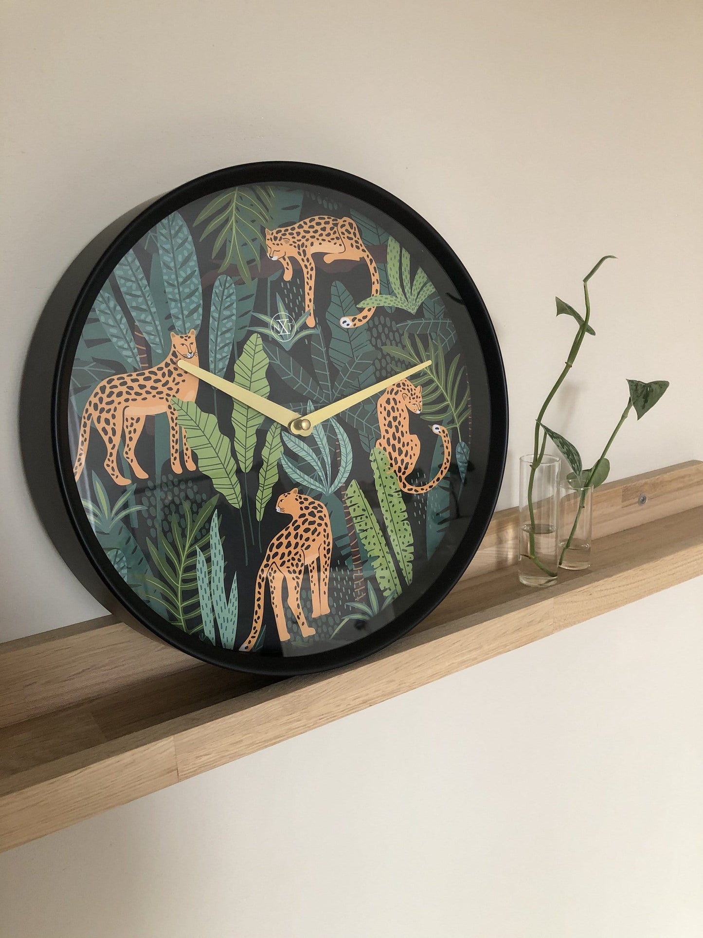 Een ronde wandklok met een zwart frame en een kleurrijk tropisch ontwerp van oranje leoparden tussen groene bladeren. Goudkleurige wijzers en een minimalistisch logo versterken de moderne, decoratieve stijl voor woonkamer of slaapkamer.