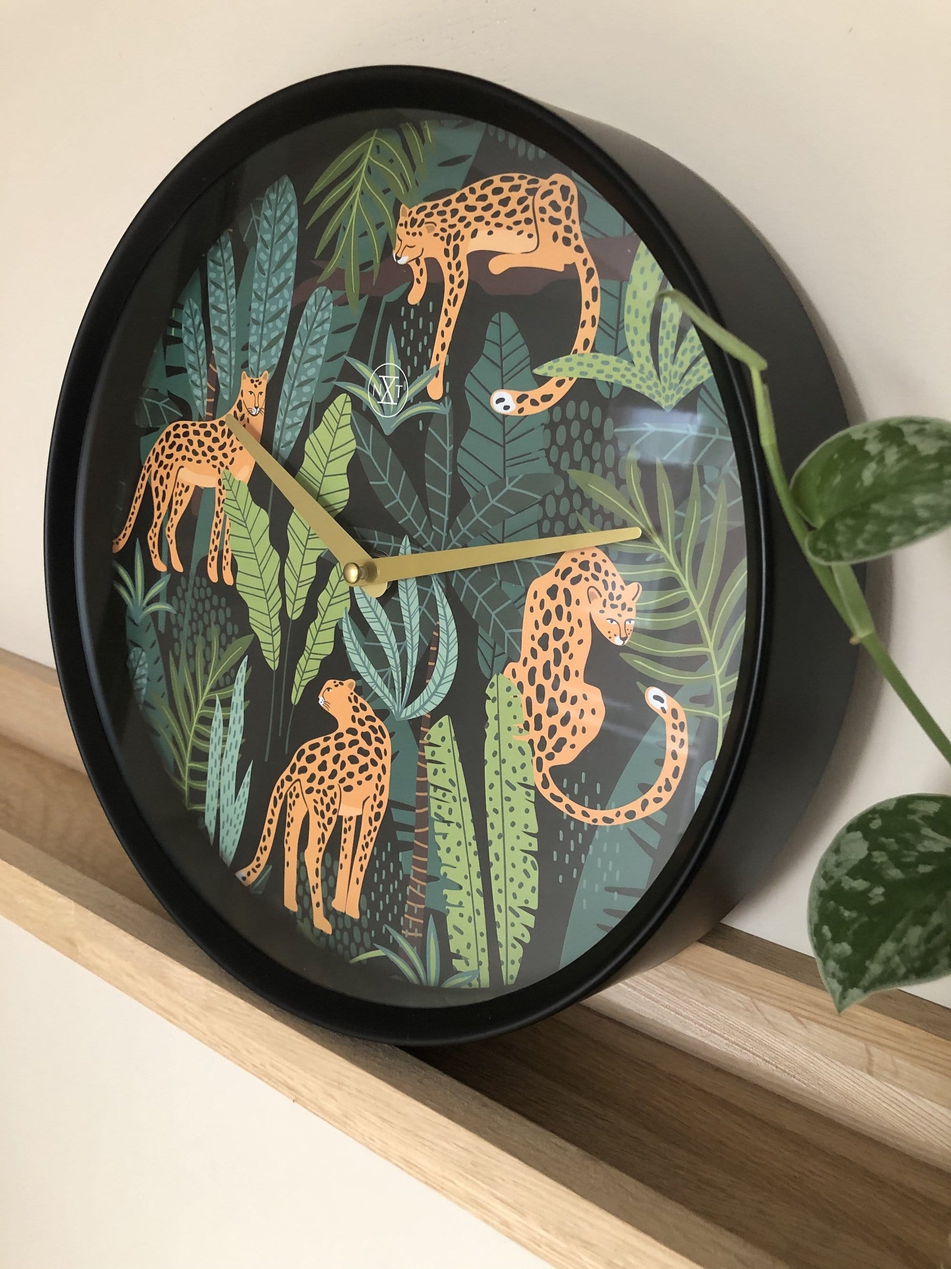 Een ronde wandklok met een zwart frame en een kleurrijk dierenmotief van oranje leoparden in een jungle. Gouden wijzers en een minimalistisch logo versterken de moderne stijl voor woonkamer of slaapkamer.