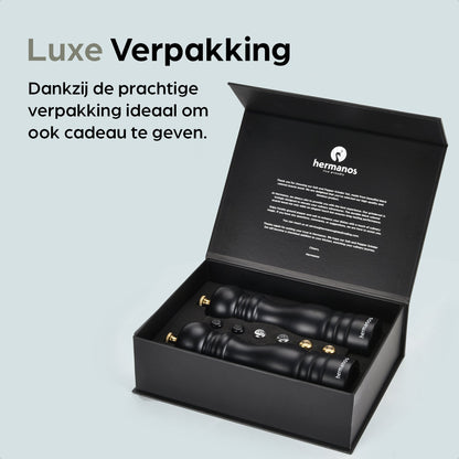 Zwarte houten pepermolen en zoutmolen met goudkleurige doppen, geplaatst op een zwarte standaard met het merk 'hermanos'. Inclusief vier vervangende doppen voor keukengebruik.