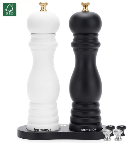 Een witte en zwarte houten pepermolen met goudkleurige doppen, geplaatst op een zwarte standaard met het merk 'hermanos'. Inclusief extra zilveren doppen voor keukengebruik.