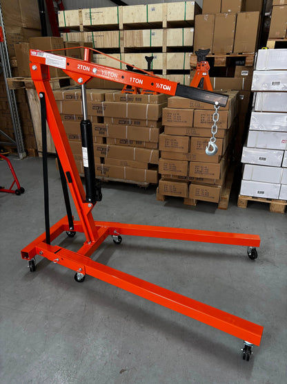 Een oranje, zwaarbelastende kraan met wielen, voorzien van een ketting en haak, staat klaar in een magazijn. Geschikt voor professioneel gebruik in werkplaatsen of op de bouwplaats.