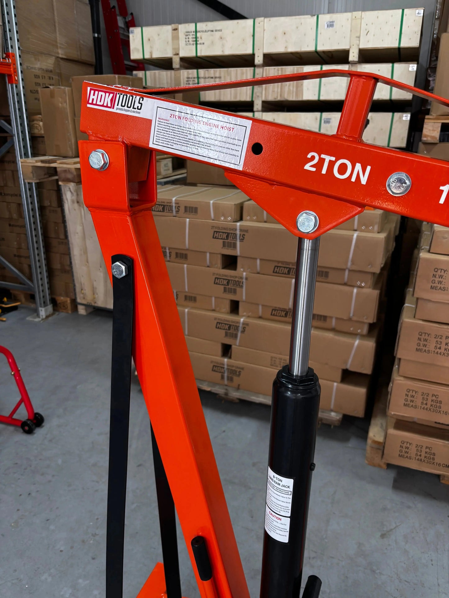 Een oranje, staafvormige kraan met wielen, uitgerust met een ketting en haak, staat klaar voor zware opdrachten in een magazijn. HDK TOOLS heavy-duty kraan voor professioneel gebruik in werkplaatsen en opslagruimtes.