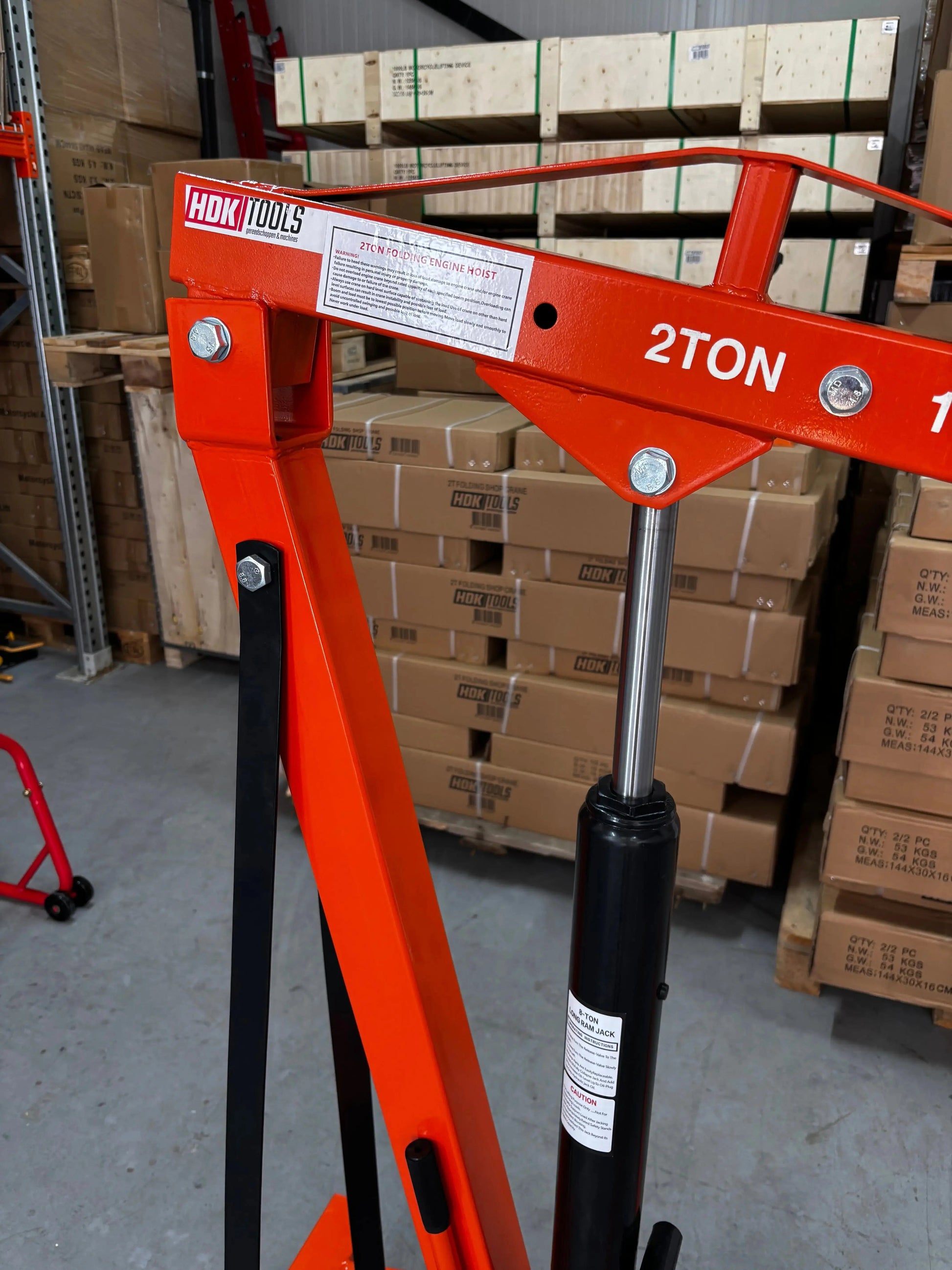 Een oranje, staafvormige kraan met wielen, uitgerust met een ketting en haak, staat klaar voor zware opdrachten in een magazijn. HDK TOOLS heavy-duty kraan voor professioneel gebruik in werkplaatsen en opslagruimtes.