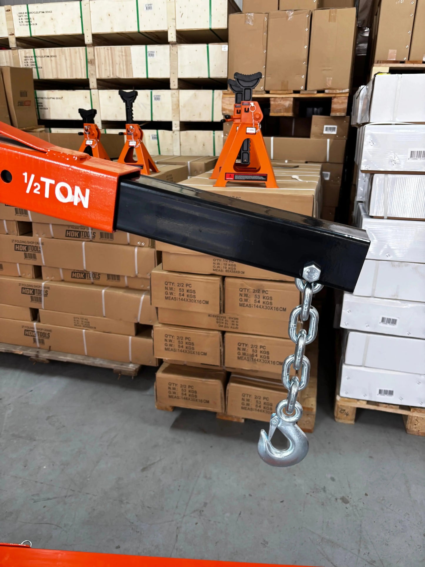 Een oranje, zwaarbelaste kraan met wielen, uitgerust met een kettinghaken en belastingaanduidingen, staat klaar voor gebruik in een industriële omgeving. Geschikt voor zware opstellingen en onderhoud in werkplaatsen of magazijnen.