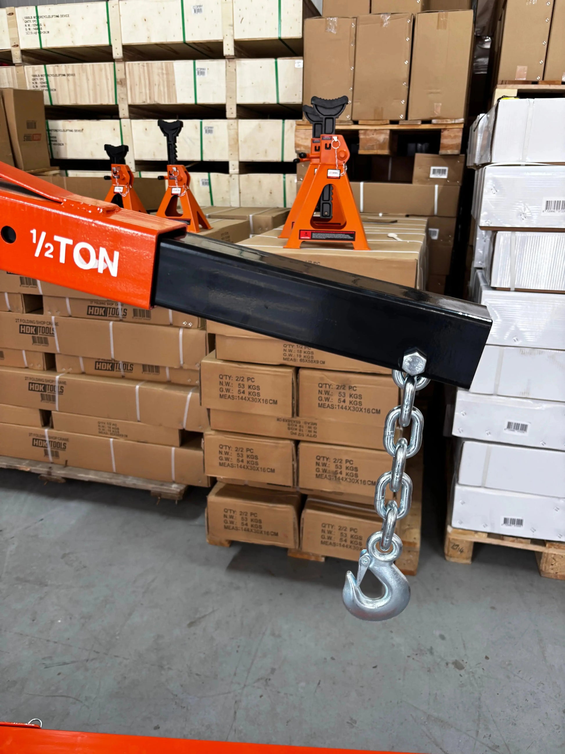 Een oranje, zwaarbelaste kraan met wielen, uitgerust met een kettinghaken en belastingaanduidingen, staat klaar voor gebruik in een industriële omgeving. Geschikt voor zware opstellingen en onderhoud in werkplaatsen of magazijnen.