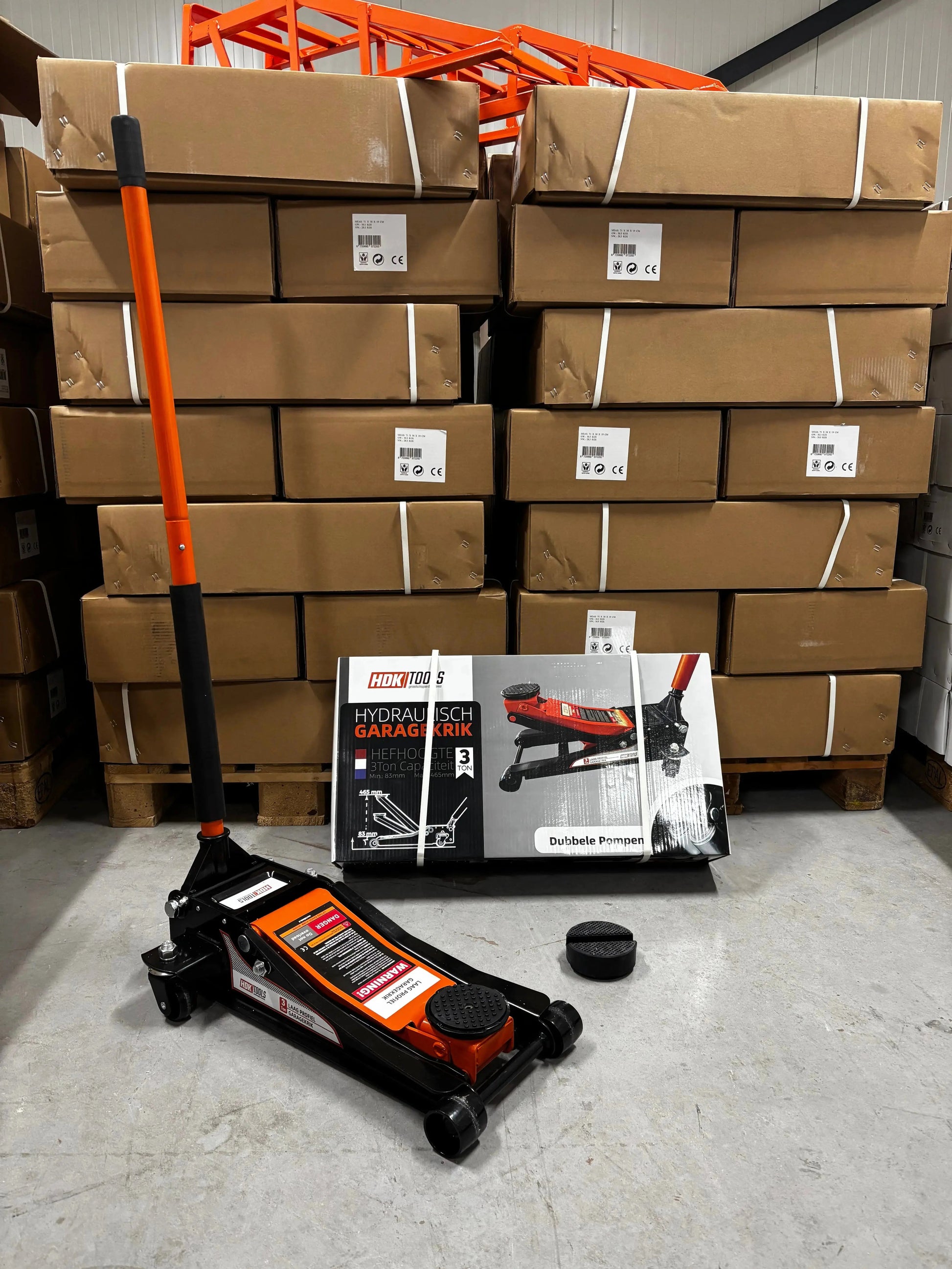 Een oranje en zwarte hydraulische garagekrik met wielen staat klaar in een professionele garage. Het apparaat is gemaakt van robuust metaal en staat naast zijn verpakking, klaar voor gebruik bij auto-onderhoud.