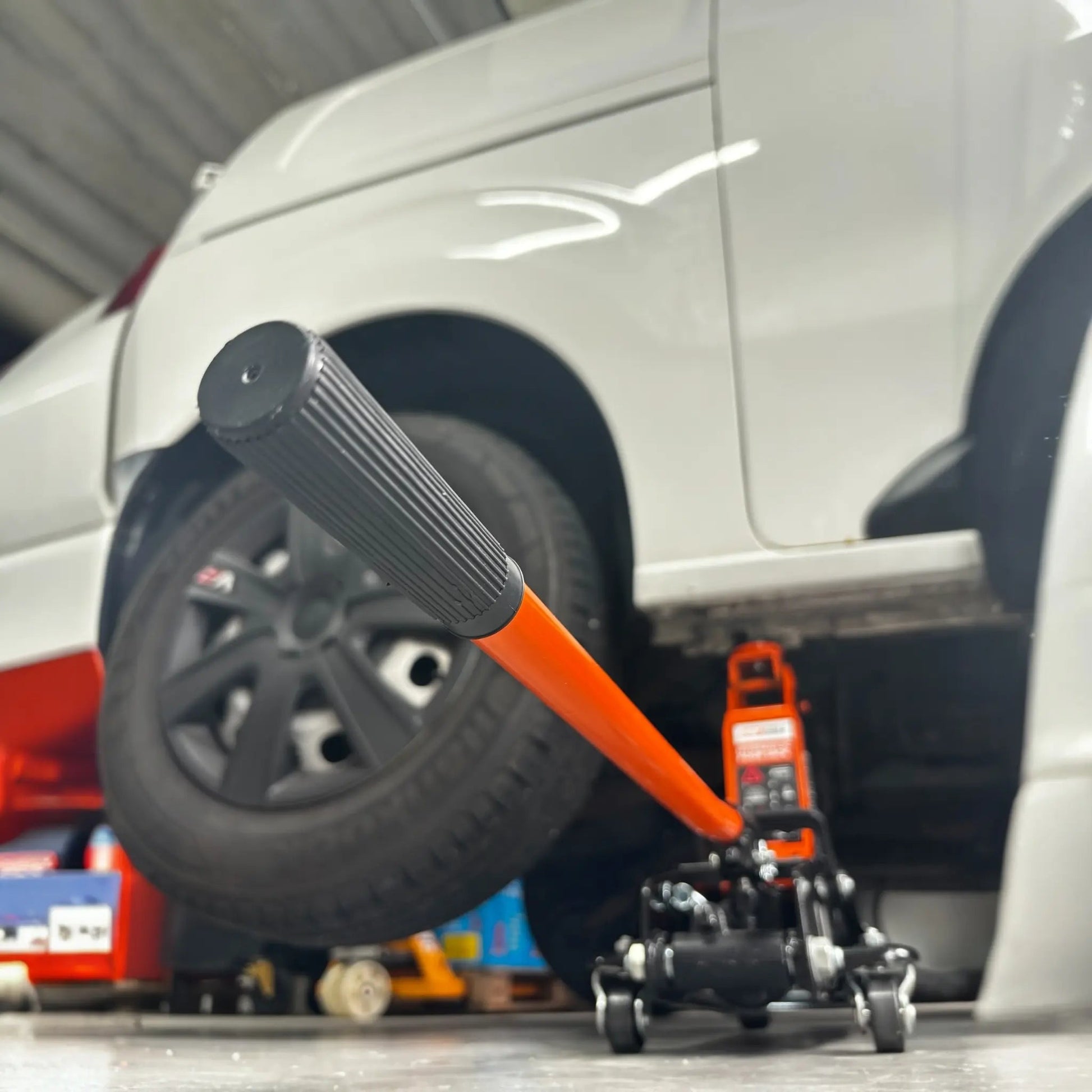 Een oranje hydraulische onderdrijf met zwarte wieltjes verhoogt een witte auto, klaar voor onderhoud of reparatie. Geschikt voor auto’s en motorfietsen, met duidelijke veiligheidsindicaties en een robuust ontwerp.