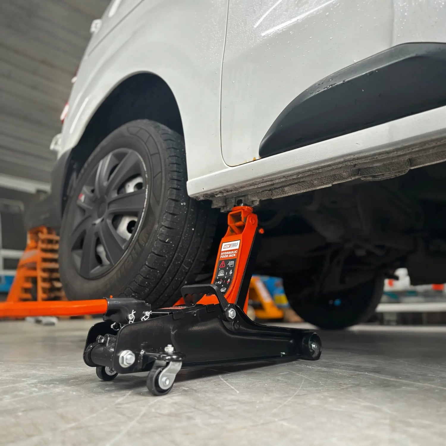 Een oranje hydraulische onderdrukpers met zwarte wieltjes verhoogt een witte auto in een garage. Geschikt voor autoreparatie en onderhoud, met duidelijke veiligheidsindicaties op het apparaat.