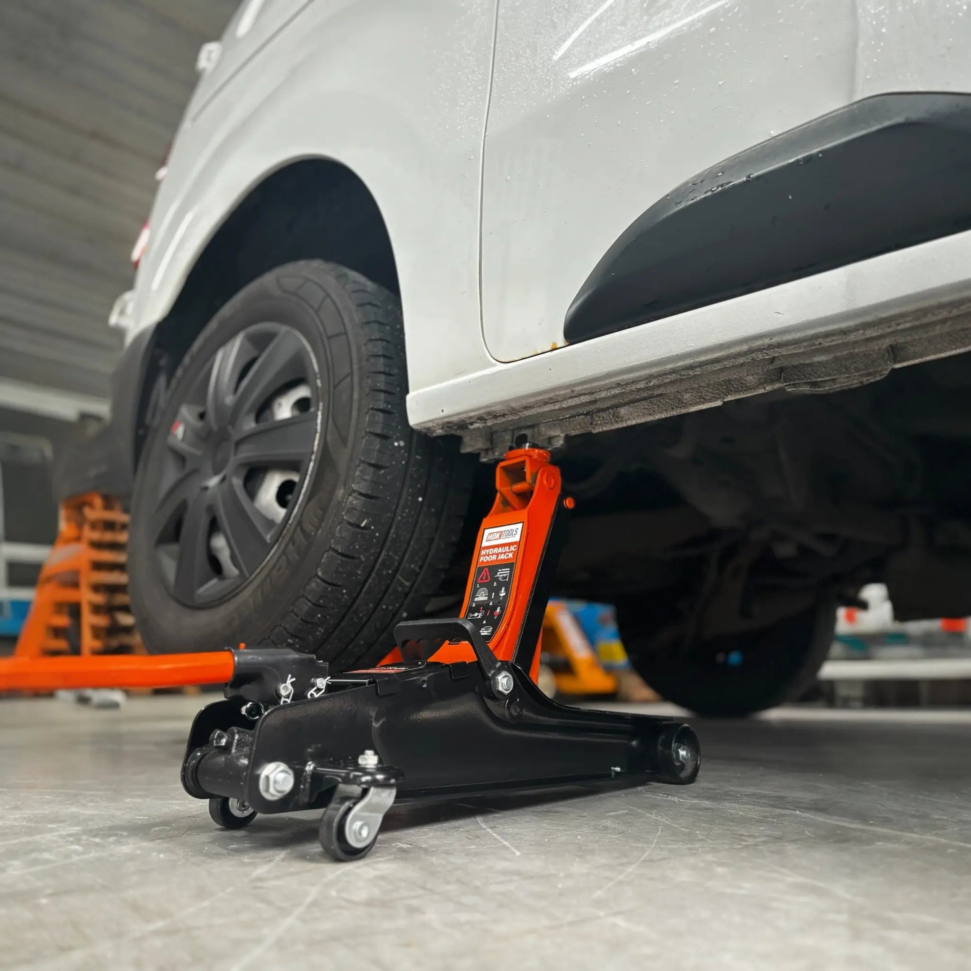 Een oranje hydraulische onderdrukpers met zwarte wieltjes verhoogt een witte auto in een garage. Geschikt voor autoreparatie en onderhoud, met duidelijke veiligheidsindicaties op het apparaat.