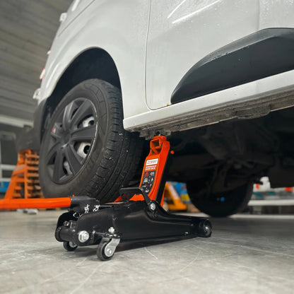 Een oranje hydraulische onderdrukpers met zwarte wieltjes verhoogt een witte auto in een garage. Geschikt voor autoreparatie en onderhoud, met duidelijke veiligheidsindicaties op het apparaat.