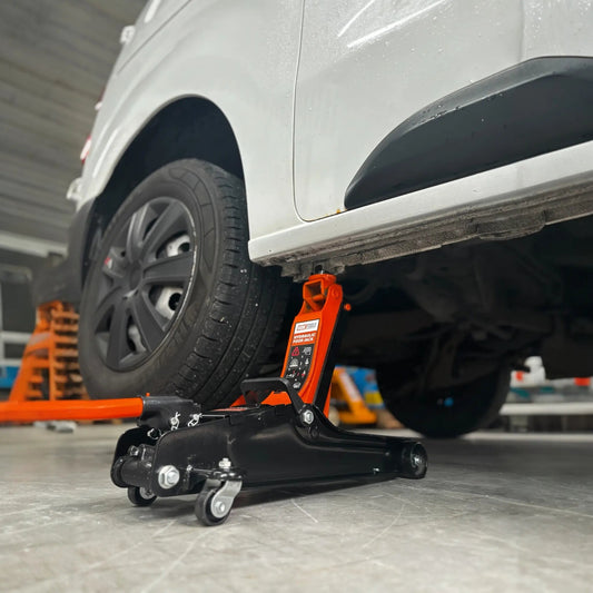 Een oranje hydraulische onderdrukpers met zwarte wieltjes verhoogt een witte auto in een garage. Geschikt voor autoreparatie en onderhoud, met duidelijke veiligheidsindicaties op het apparaat.