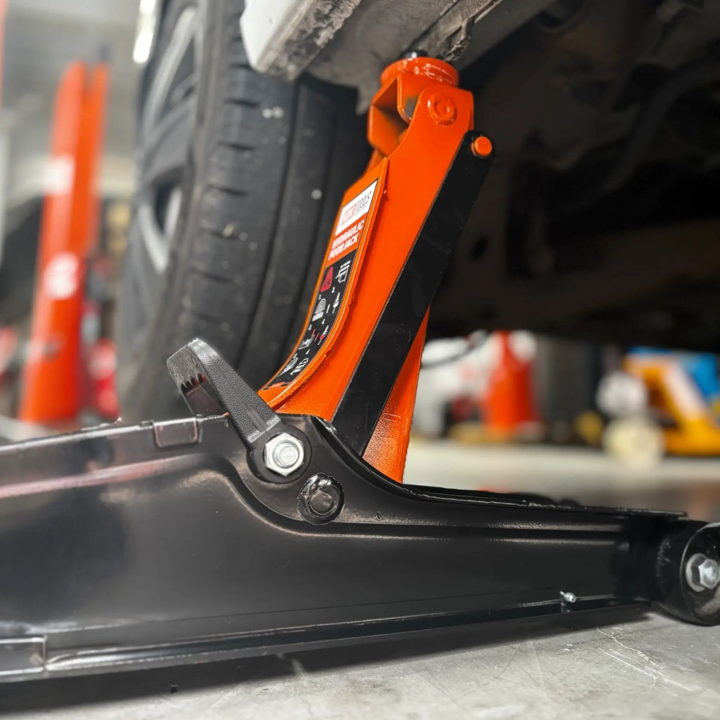 Een oranje hydraulische onderdrukpers met zwarte wielen verhoogt een witte auto in een garage. Geschikt voor auto-onderhoud en reparatie, met duidelijke veiligheidsindicaties op het apparaat.