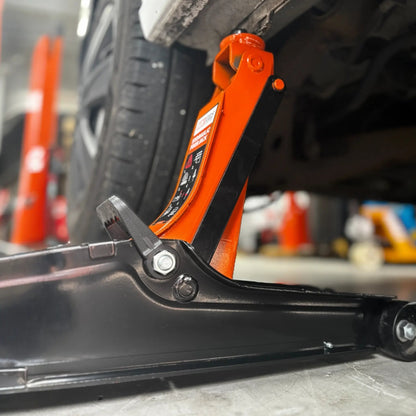 Een oranje hydraulische onderdrukpers met zwarte wielen verhoogt een witte auto in een garage. Geschikt voor auto-onderhoud en reparatie, met duidelijke veiligheidsindicaties op het apparaat.