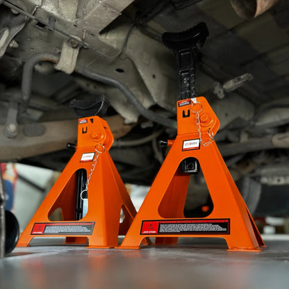 Twee oranje, 6-ton jackstands onder een auto, met zwarte handgrepen en waarschuwingslabels. Geschikt voor veilig onderhoud en reparatie van voertuigen in de garage.