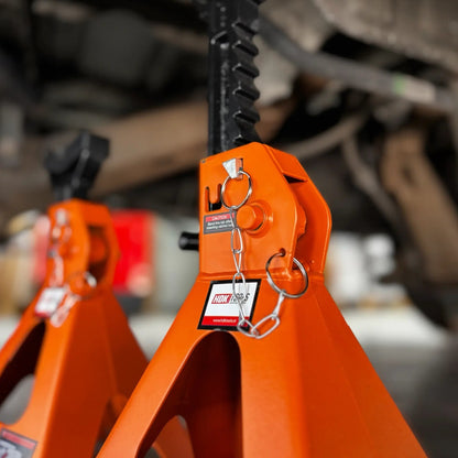 Twee oranje, 6-ton jackstands onder een auto, met zwarte handgrepen en waarschuwingslabels. Geschikt voor veilig onderhoud en reparatie van voertuigen in de garage.