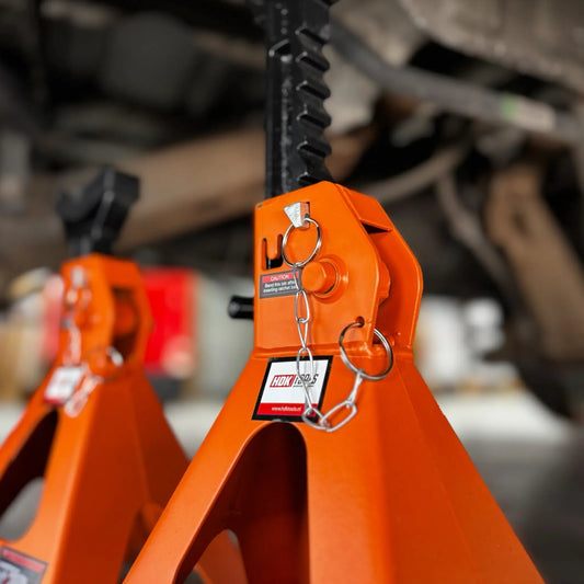Twee oranje, 6-ton jackstands onder een auto, met zwarte handgrepen en waarschuwingslabels. Geschikt voor veilig onderhoud en reparatie van voertuigen in de garage.