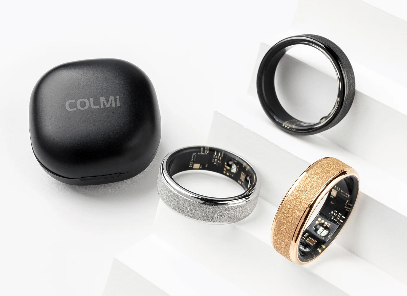 Een COLMi slimme ring in goudkleur met glinsterende textuur, naast een zwarte variant met zichtbare technische componenten, beide in een zwarte opbergdoos voor dagelijks gebruik en sportactiviteiten. - Foto 32