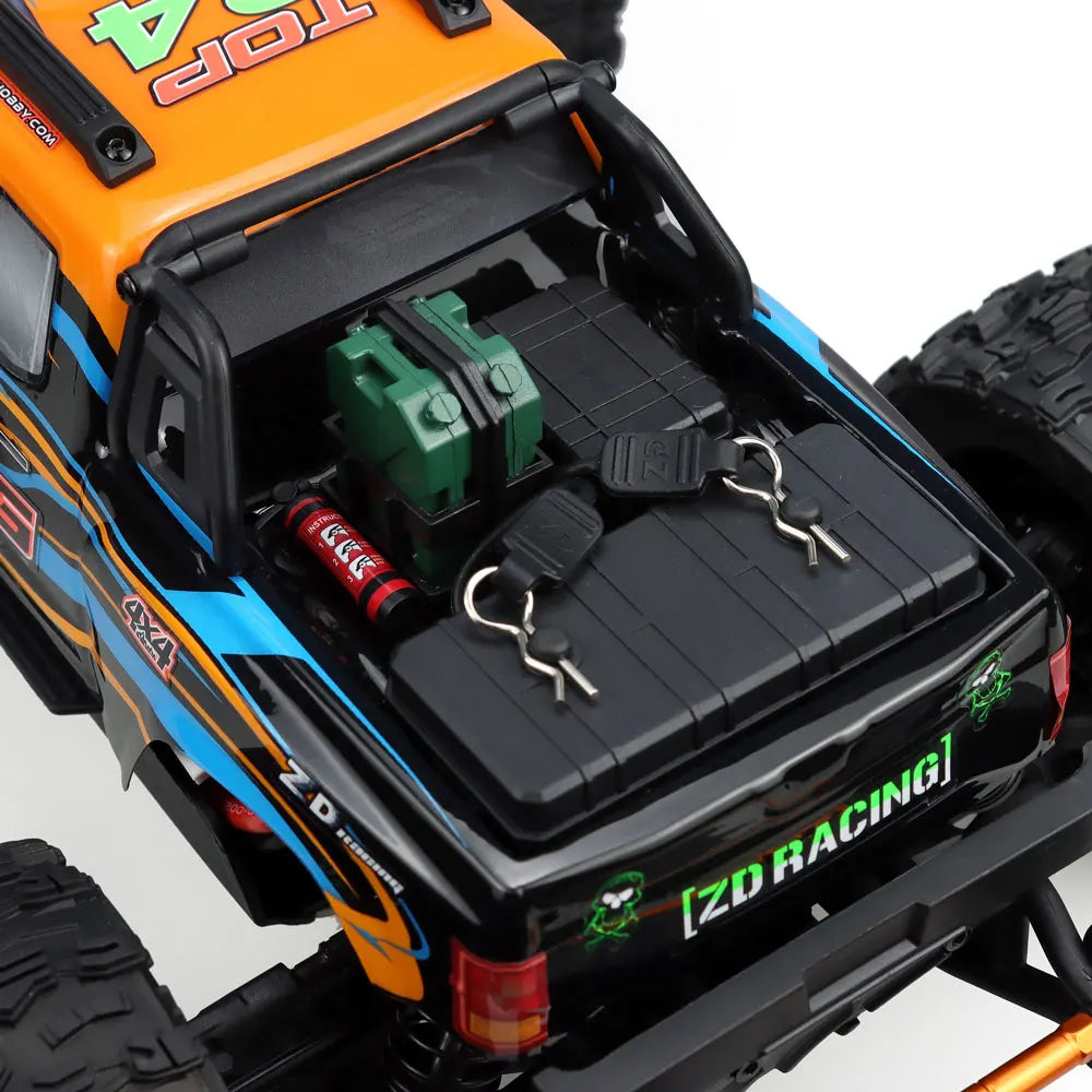 Oranje en blauwe RC monstertruck met grote zwarte banden en zichtbare "MT-16" sticker, geplaatst naast zijn zwarte stuurunit voor terreinrijden en spe