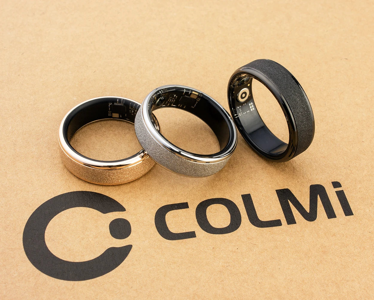 Een COLMi slimme ring in goudkleur met glinsterend oppervlak, naast een zwarte variant met zichtbare technische componenten, beide in een zwarte opbergdoos voor dagelijks gebruik en sportactiviteiten. - Foto 30