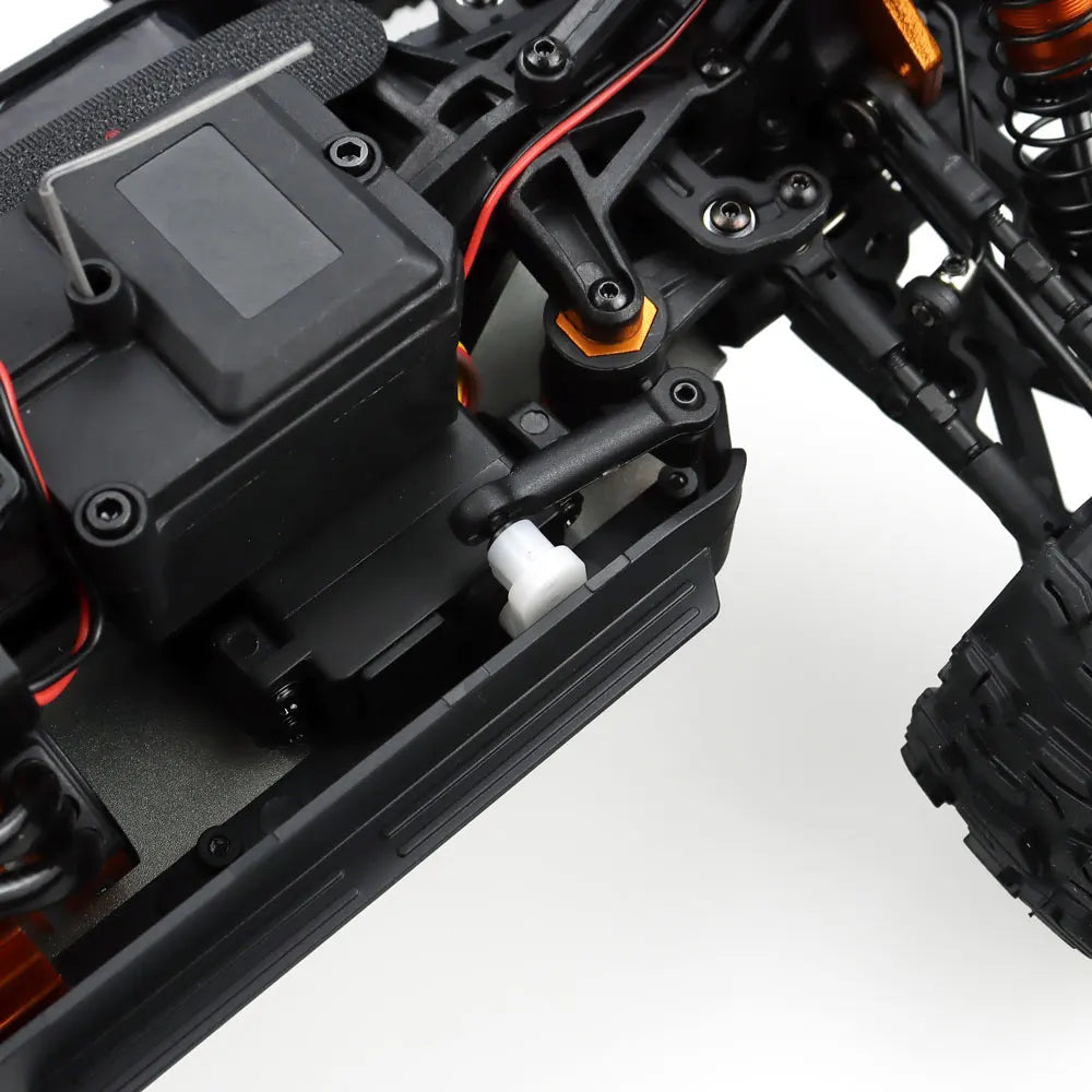 Oranje en blauwe RC monstertruck met grote zwarte banden en zichtbare "MT-16" sticker, geplaatst naast zijn zwarte stuurunit voor speelplezier op zand