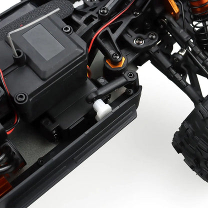 Oranje en blauwe RC monstertruck met grote zwarte banden en zichtbare "MT-16" sticker, geplaatst naast zijn zwarte stuurunit voor speelplezier op zand