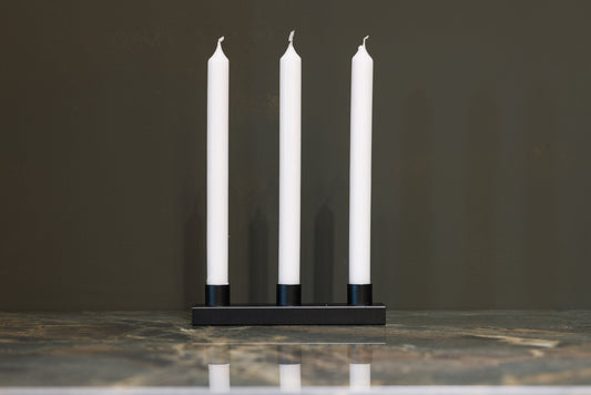 Een minimalistisch zwart kaarsenhouder met drie witte kaarsen, geplaatst op een marmeren oppervlak. Perfect voor een moderne, rustige sfeer in woon- of eetkamer.