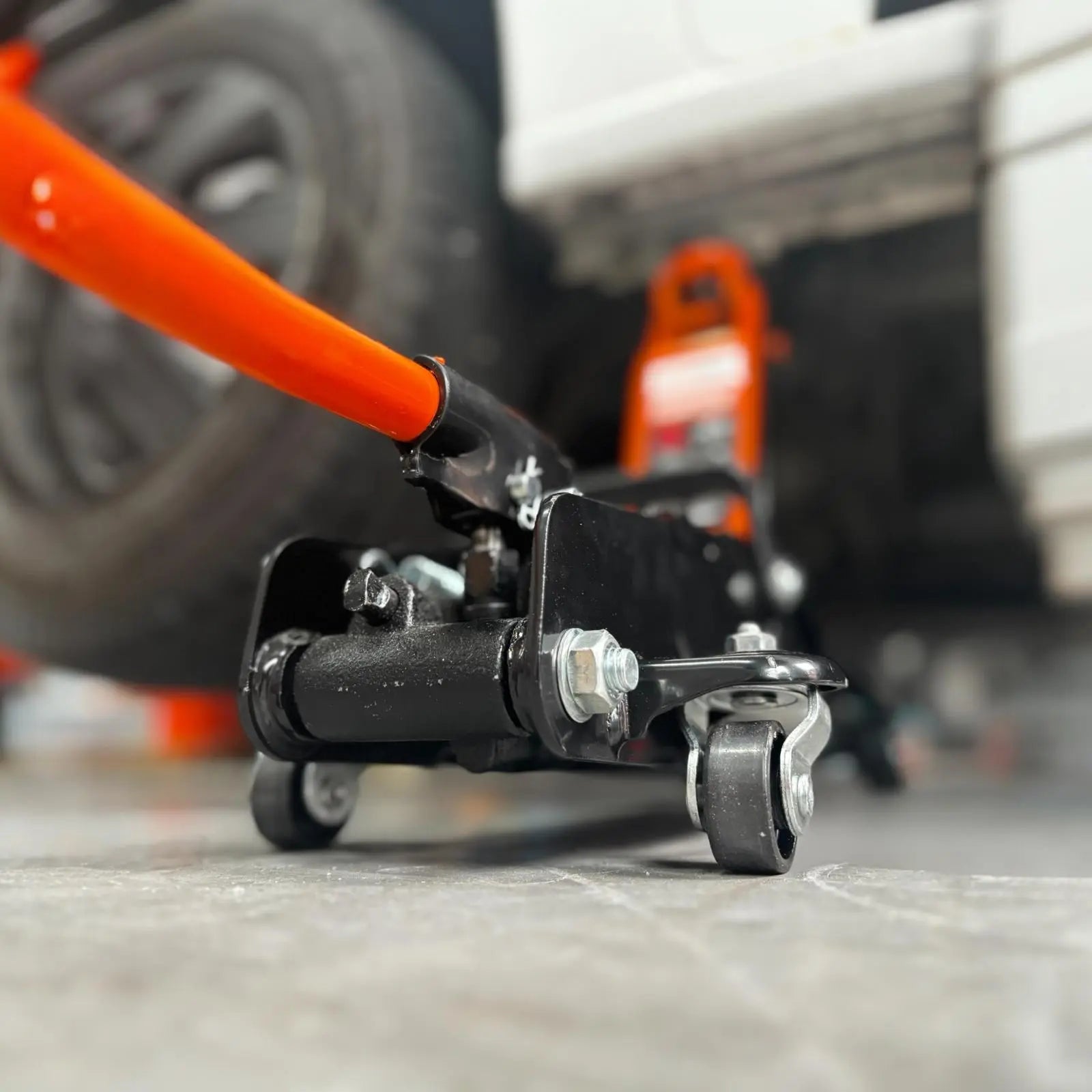 Een oranje hydraulische onderdrukpers met zwarte wielen ondersteunt een witte auto in een garage. Geschikt voor auto-onderhoud en reparaties, met duidelijke veiligheidsindicaties op het apparaat.